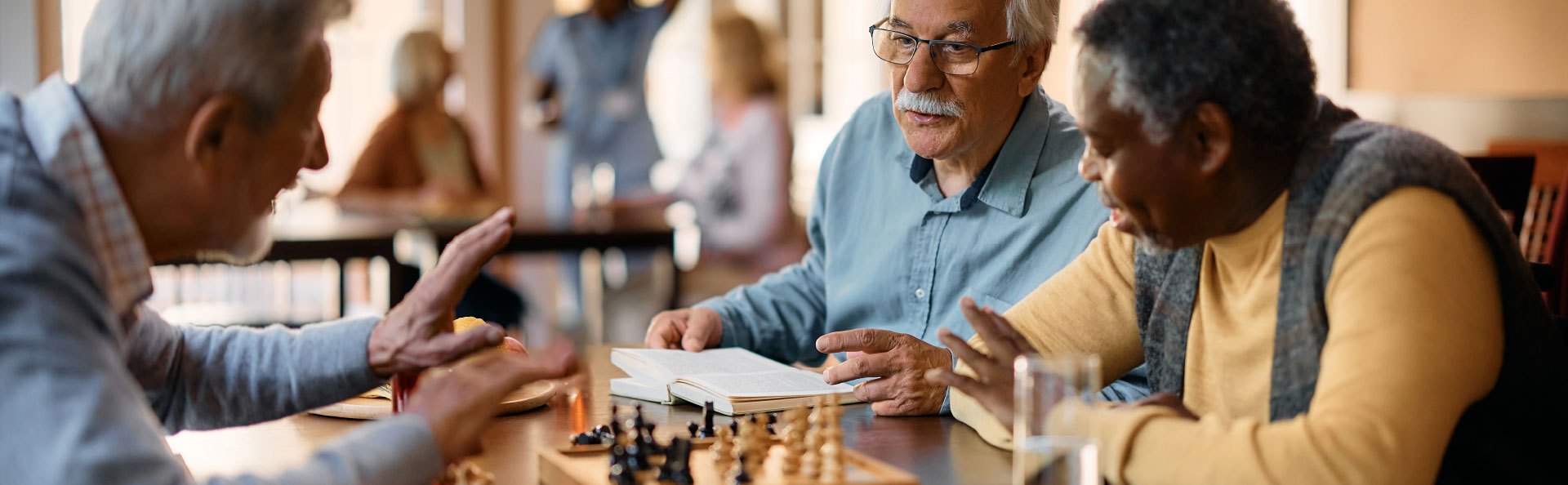 Three seniors site at a table with a chess set and books.